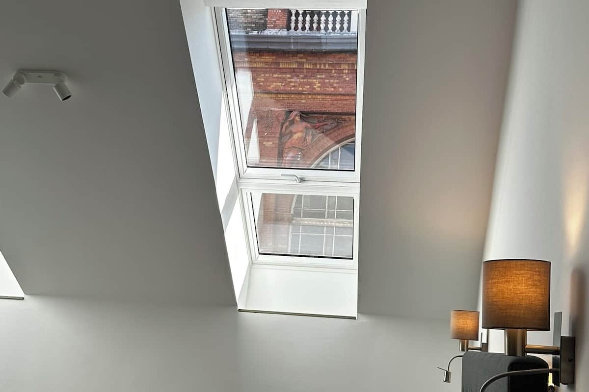 A hotel room with a bed, a red blanket, a nightstand with a lamp, and a view of a building through a skylight.
