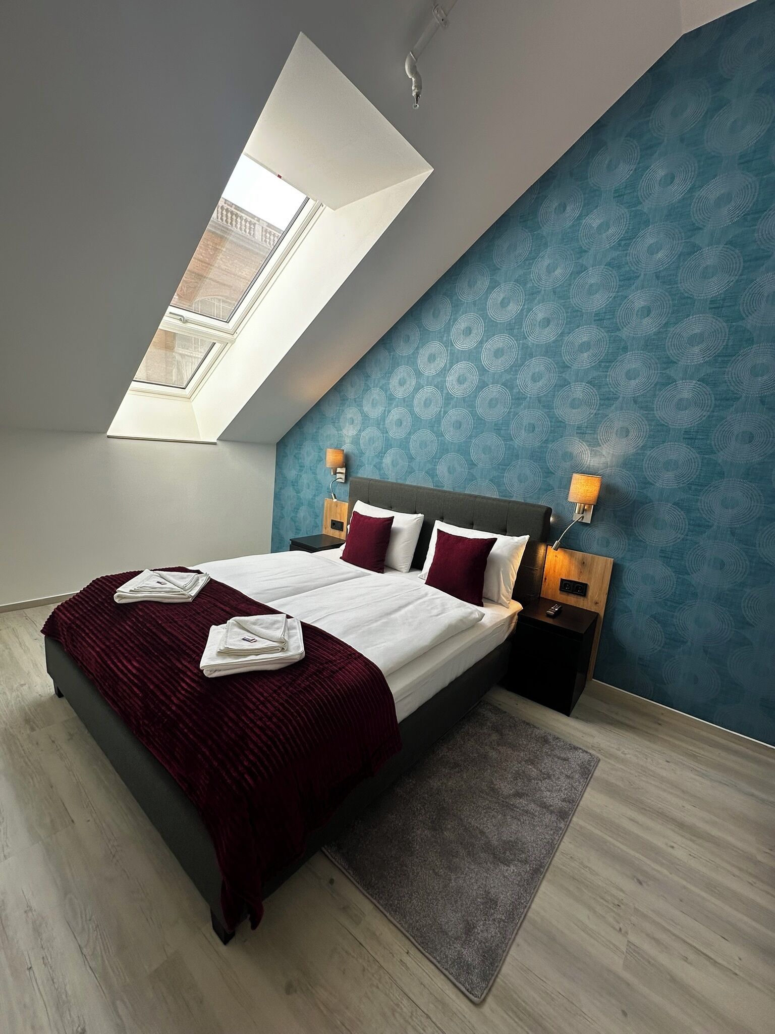A modern bedroom with a skylight, a bed with a red throw, two bedside lamps, and a patterned wall.