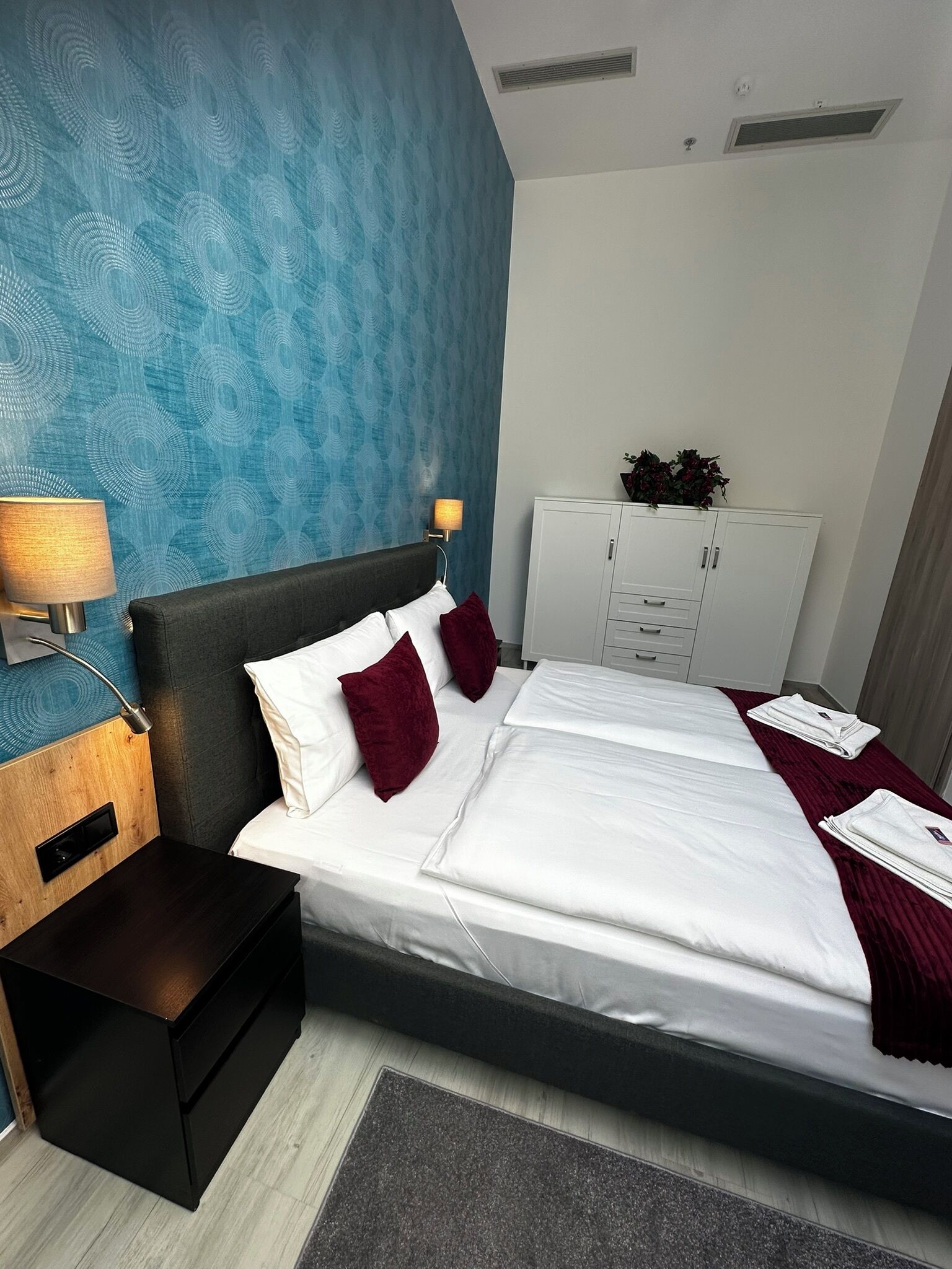 Une chambre d’hôtel avec un lit, une table de chevet, une lampe, une armoire et un mur orné d’un motif bleu.
