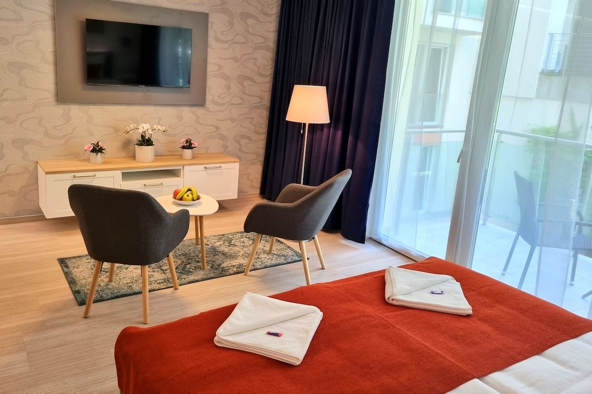 A hotel room with a bed, two chairs, a desk with a TV, a lamp, and a view of the outdoors.