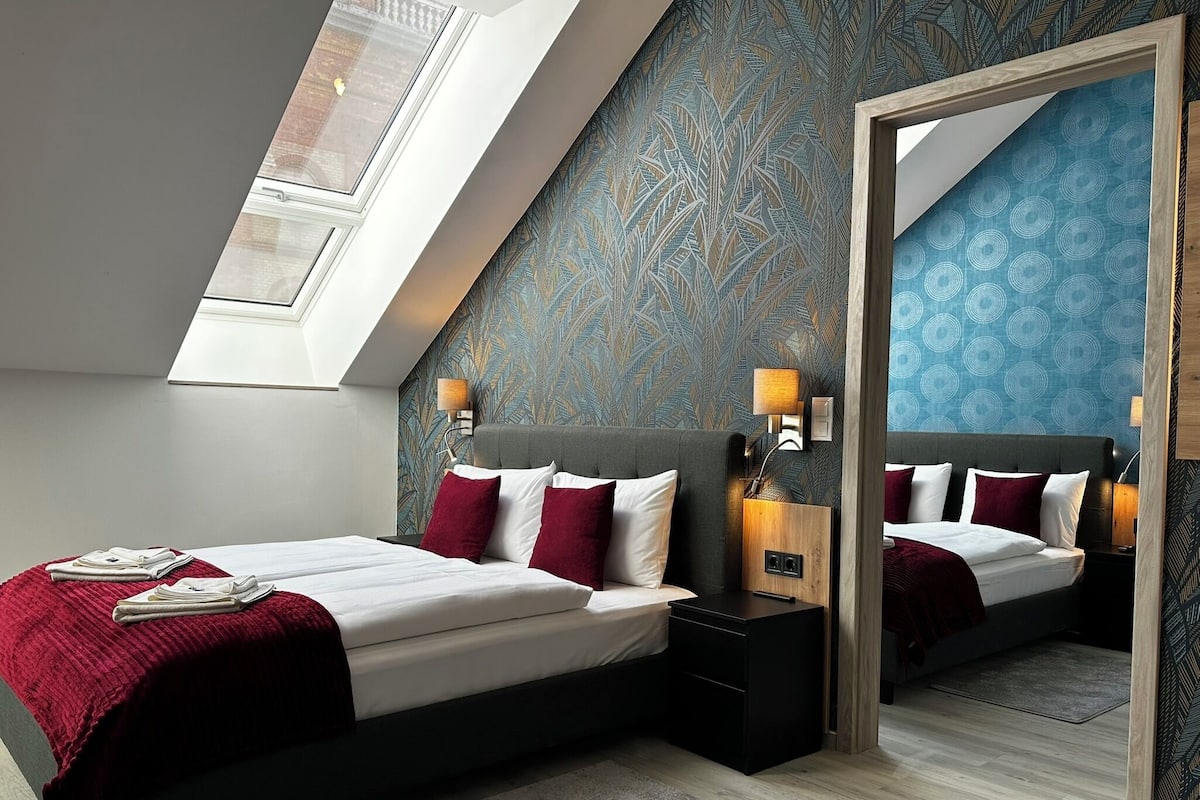 A modern bedroom with a skylight, a bed with red and white bedding, a nightstand, and a mirror.