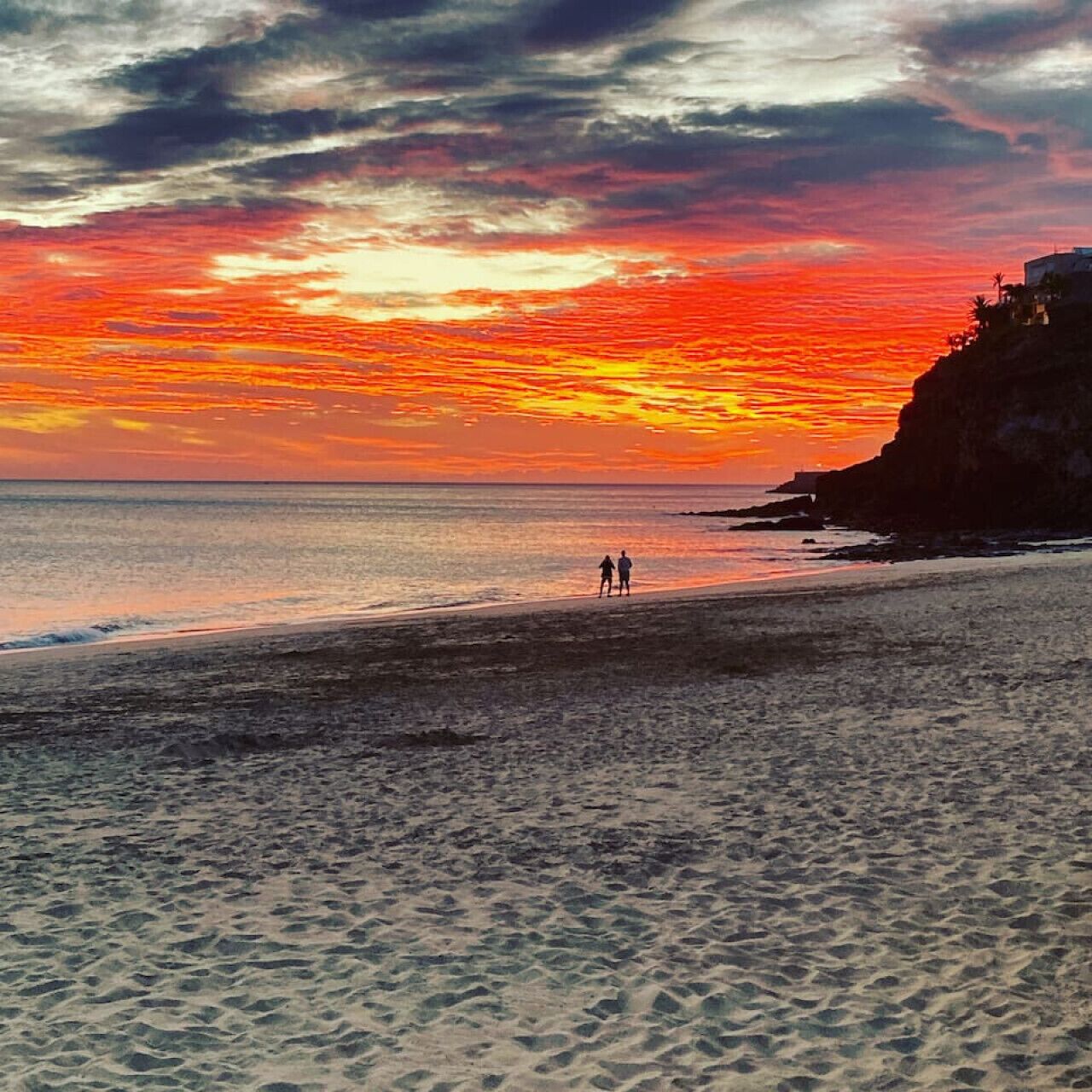 A beach with two people walking along the shore, a cliff on the right, and a sunset in the background.