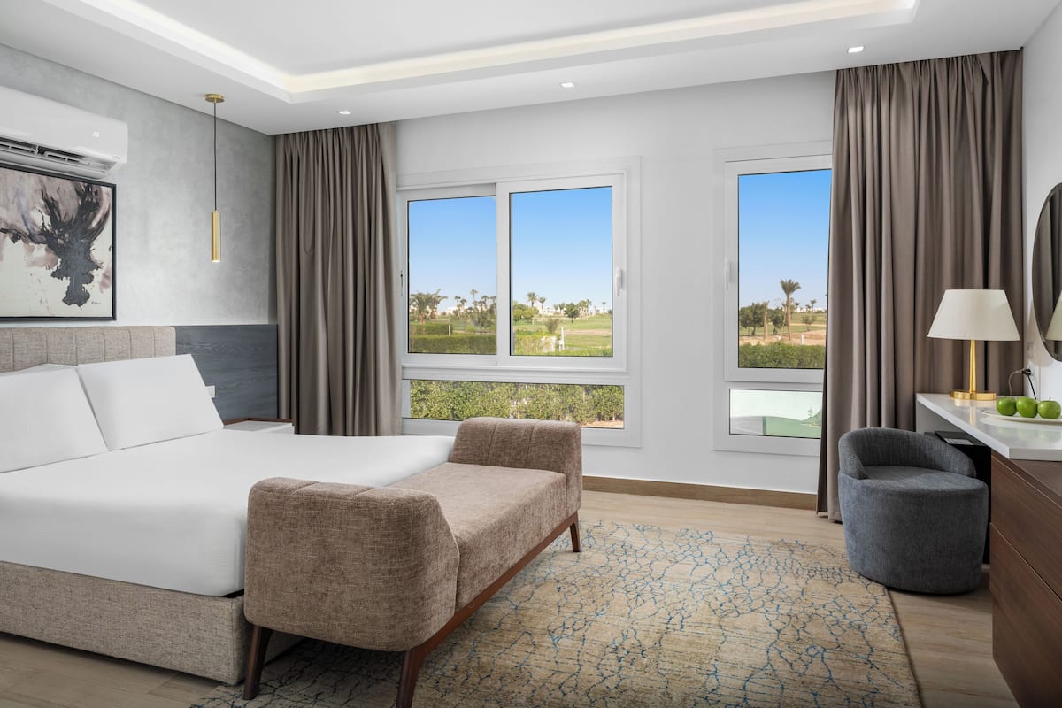 Camera d'albergo moderna con un letto grande, due poltrone, una scrivania con una lampada e vista su un campo da golf.