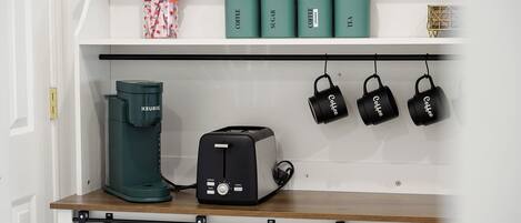 A kitchen shelf with a coffee maker, sugar, coffee, and tea containers, and hanging coffee mugs.