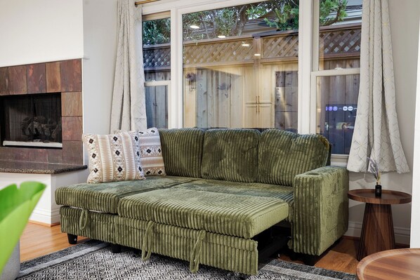 A green sofa with a patterned cushion, a fireplace, and a view of a wooden fence through a window. - LakeFront Lavender House (San Mateo)