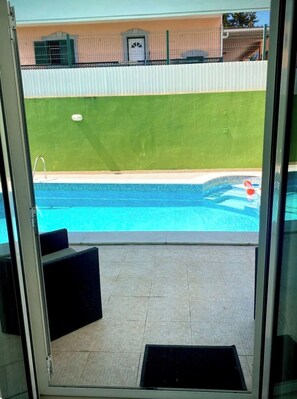 A view through a glass door of a swimming pool with a tiled deck, a bench, and a building with a green wall and a fence in the background.