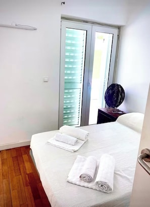 A hotel room with a bed, two rolled towels, a nightstand with a lamp, and a window with blinds.