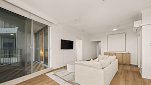 A modern living room with a large sliding glass door leading to a balcony, a flat-screen TV mounted on the wall, and a wooden floor. - Absolute Beachfront Apartment with Direct Pool & Beach Access (Main Beach)