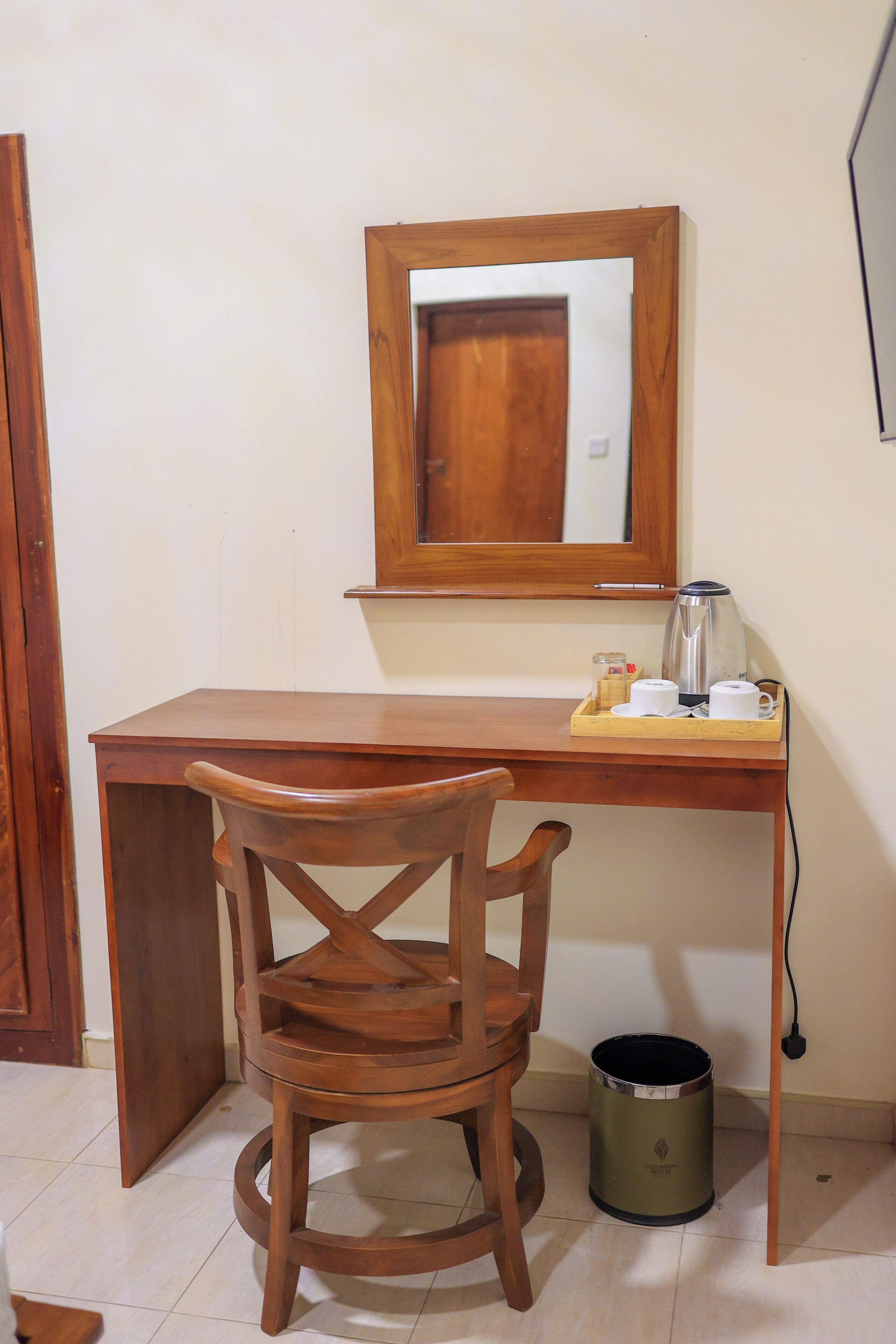 A wooden desk with a chair, a mirror, a kettle, and a trash can.