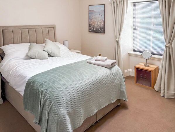 A neatly made bed with a light-colored headboard, a bedside table with a lamp, and a window with curtains. - 4 bedroom accommodation in Inverkip, near Greenock (Inverkip, near Greenock)