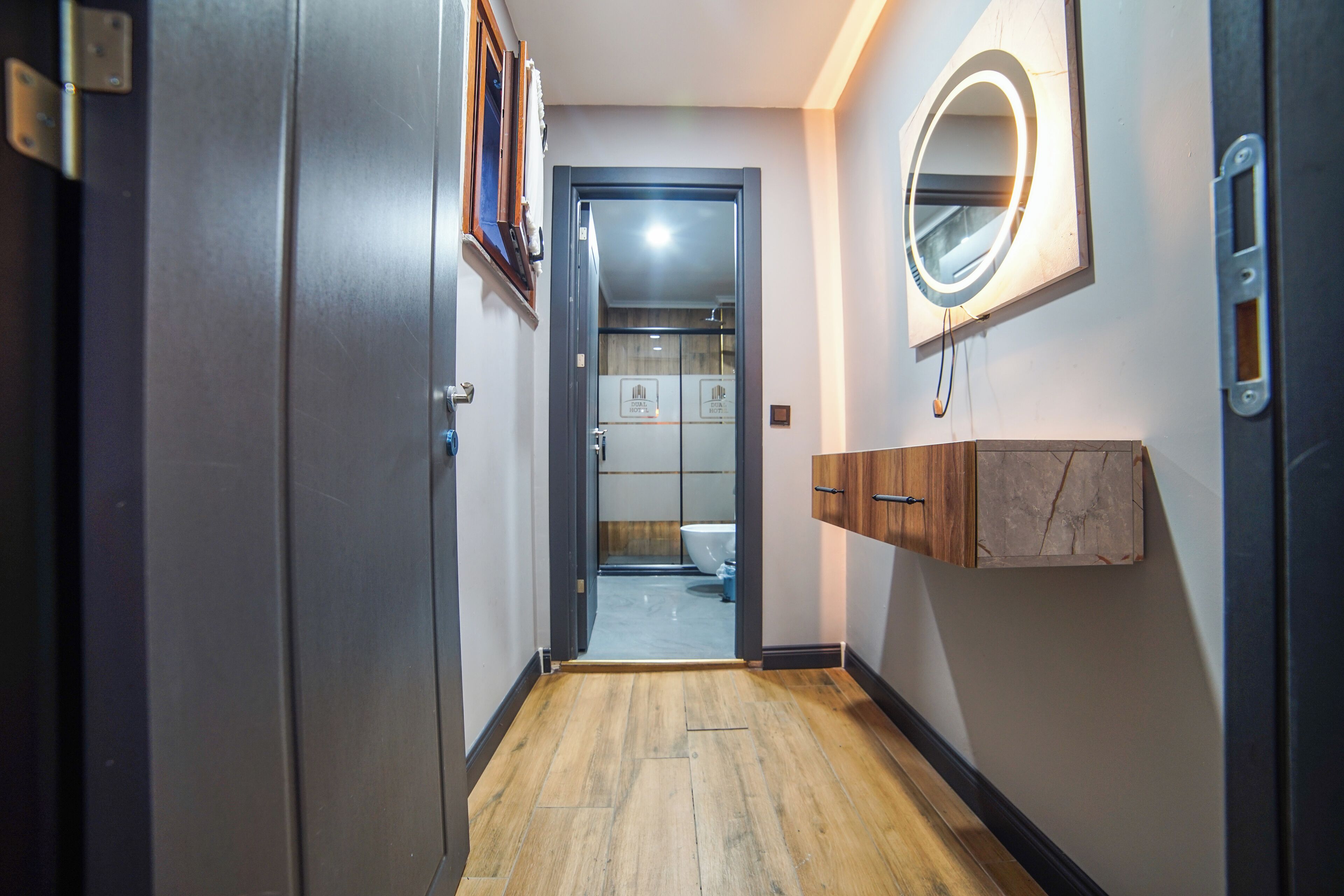 A hallway with a wooden floor, a door leading to a bathroom, a mirror with a light, and a wooden cabinet.