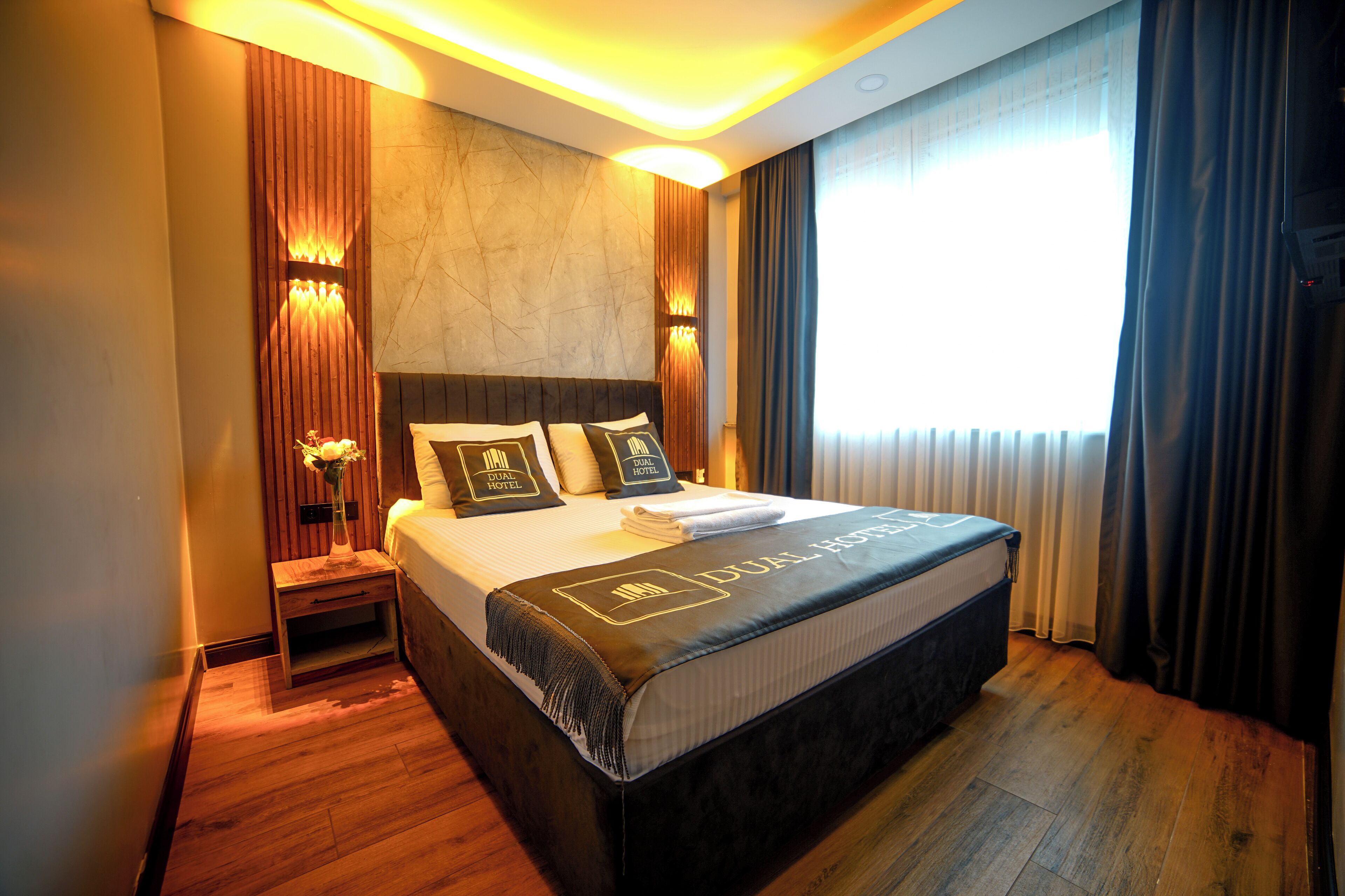 Une chambre d’hôtel avec un grand lit, une table de chevet, un vase avec des fleurs et un luminaire fixé au mur.