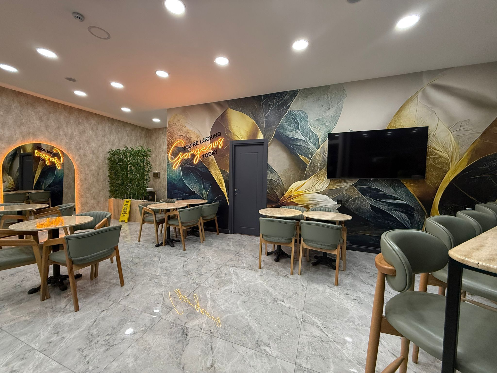 A modern restaurant interior with marble flooring, green upholstered chairs, and a large mural with leaf patterns.
