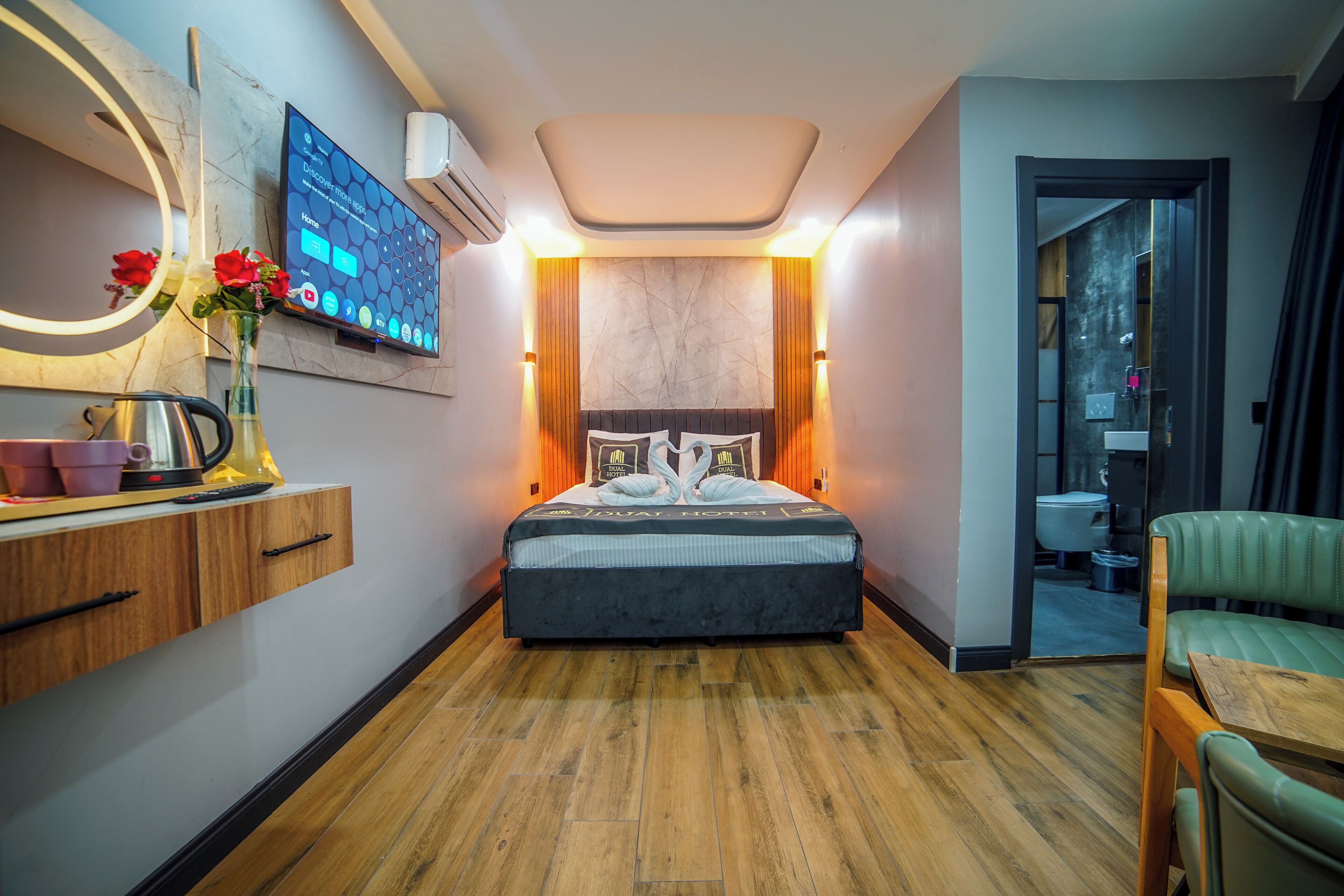 A modern hotel room with a large bed, a bedside table with a vase of flowers, a wall-mounted TV, and a wooden floor.