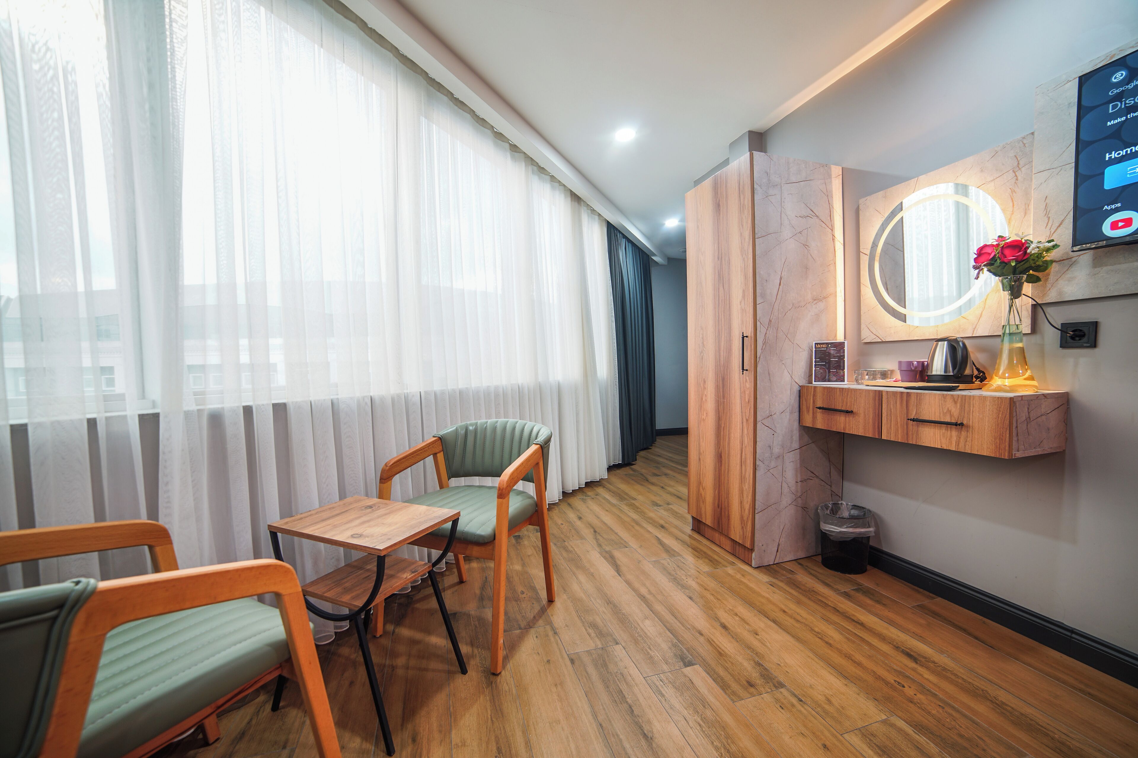 A hotel room with a wooden floor, a green upholstered chair, a wooden table, a small wooden cabinet with a mirror, and a window with sheer curtains.