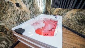 Un baño moderno con pared de mármol, una bañera blanca y sales de baño rojas, y suelo de madera.