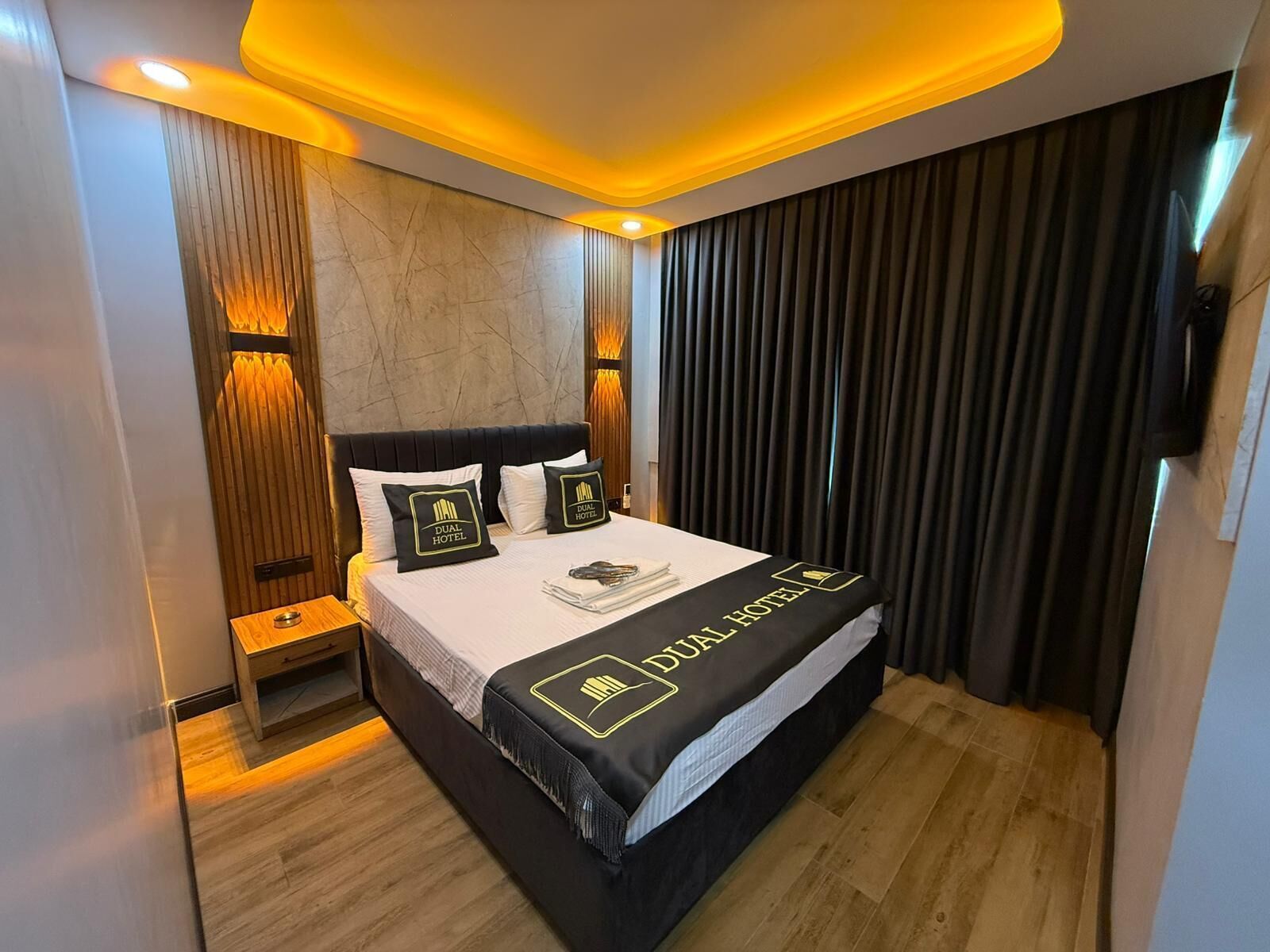 A modern hotel room with a large bed, a nightstand, a wooden panel wall, and a dark curtain.