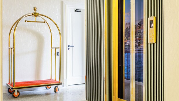 A hallway with a red and gold luggage cart, a white door with a 'Laundry' sign, and a glass elevator with a cityscape view. - Marmaris Saray Hotel (Tashkent)