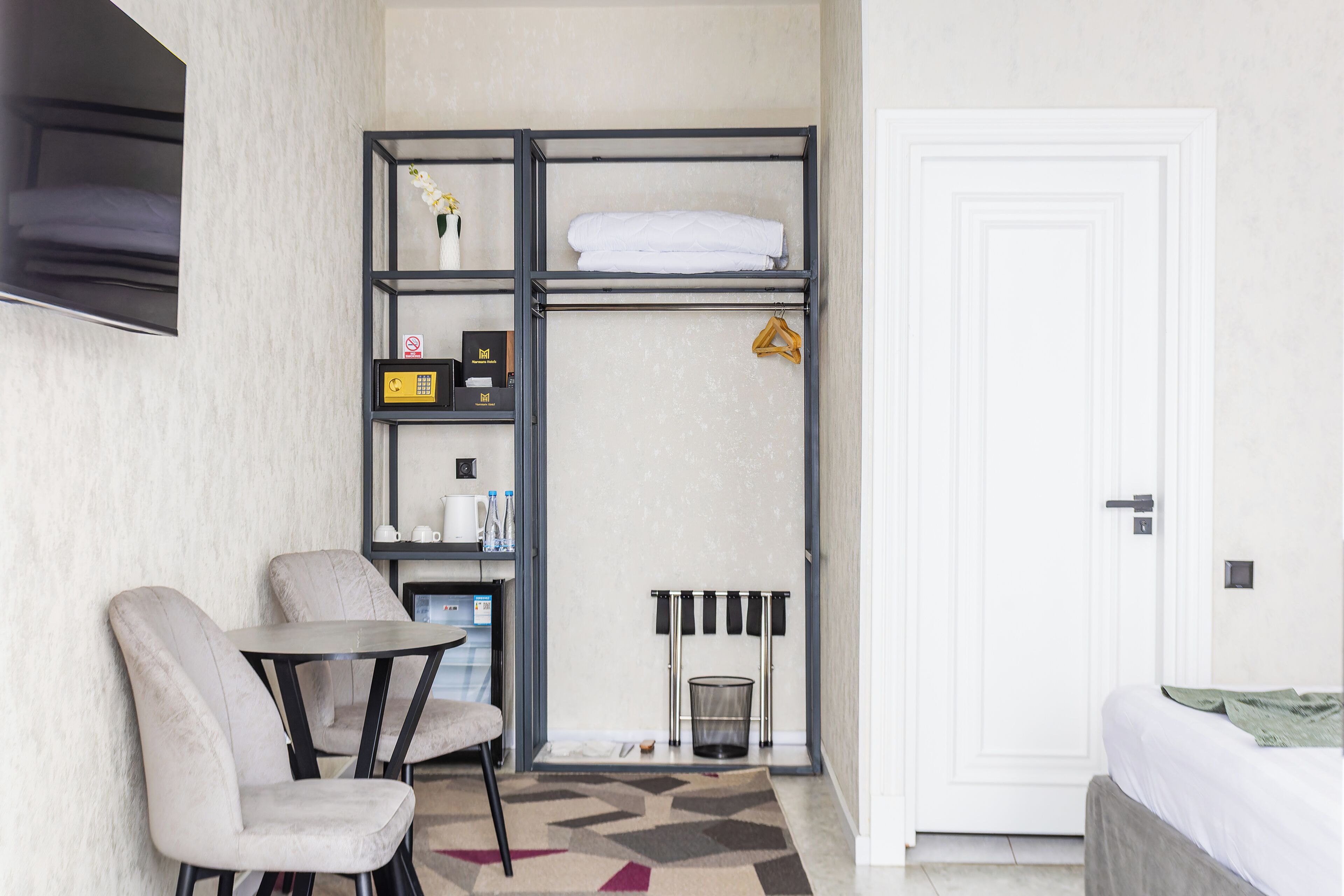 Quarto de hotel compacto com cama, área de estar com mesa e cadeiras, nicho embutido com artigos de higiene e uma porta.