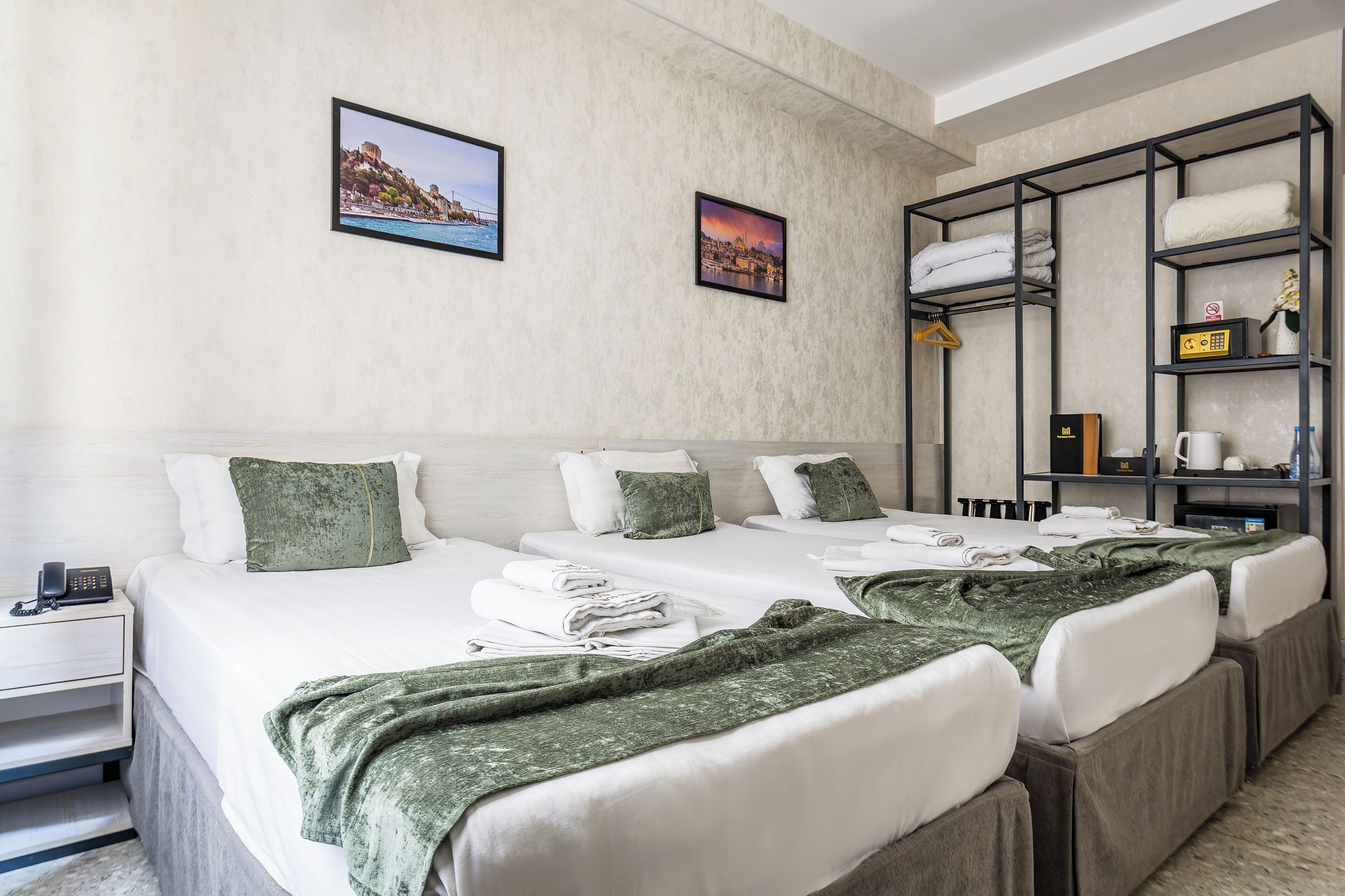 A hotel room with two beds, a bedside table with a phone, a shelf with towels and toiletries, and two framed pictures on the wall.