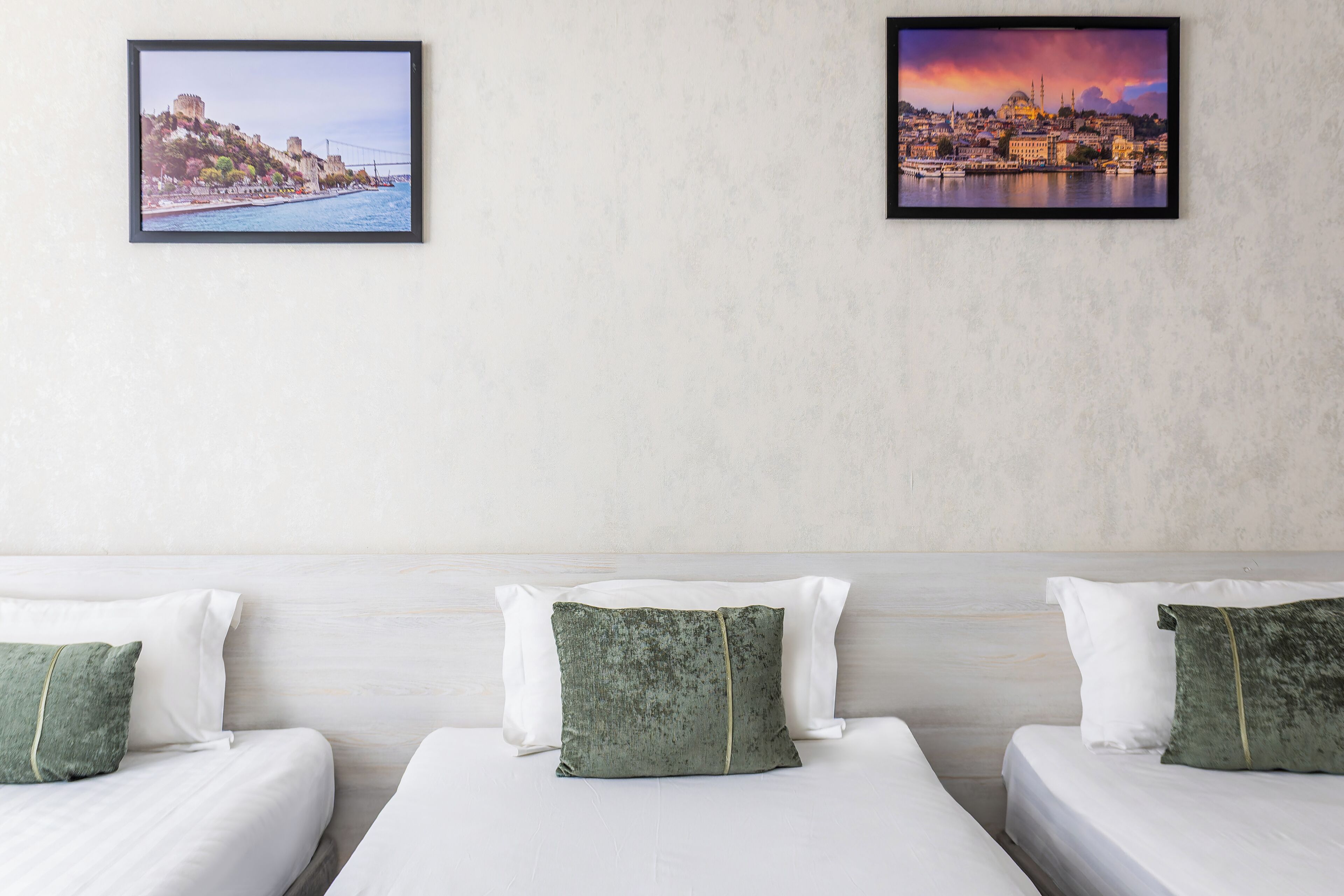 A hotel room with two beds, each with white linens and green pillows. There are two framed pictures on the wall, one depicting a coastal cityscape and the other a sunset cityscape.