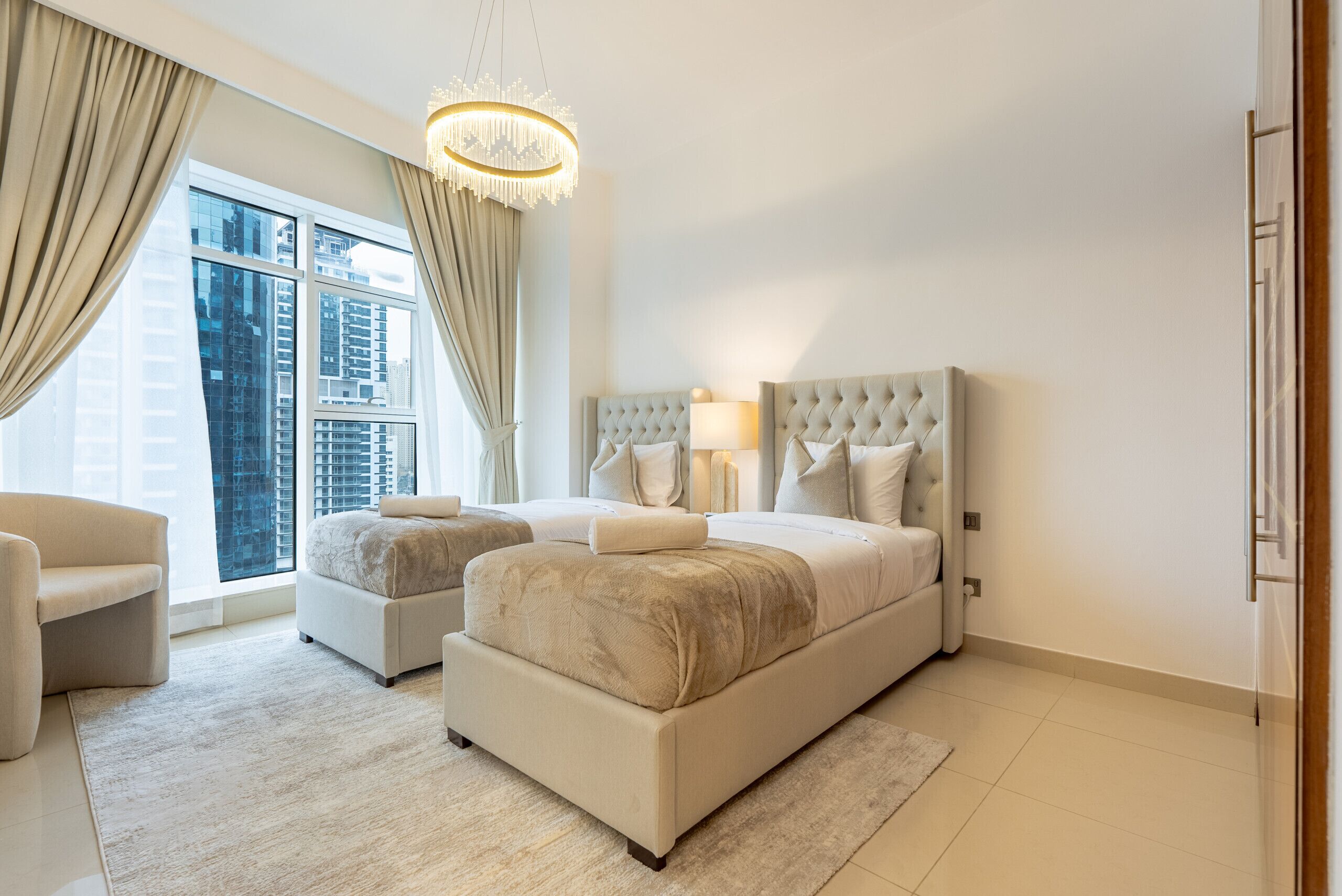 Ein modernes Schlafzimmer mit einem großen Bett, zwei Sessel, einer Kandelaber und Blick auf hohe Gebäude durch große Fenster.