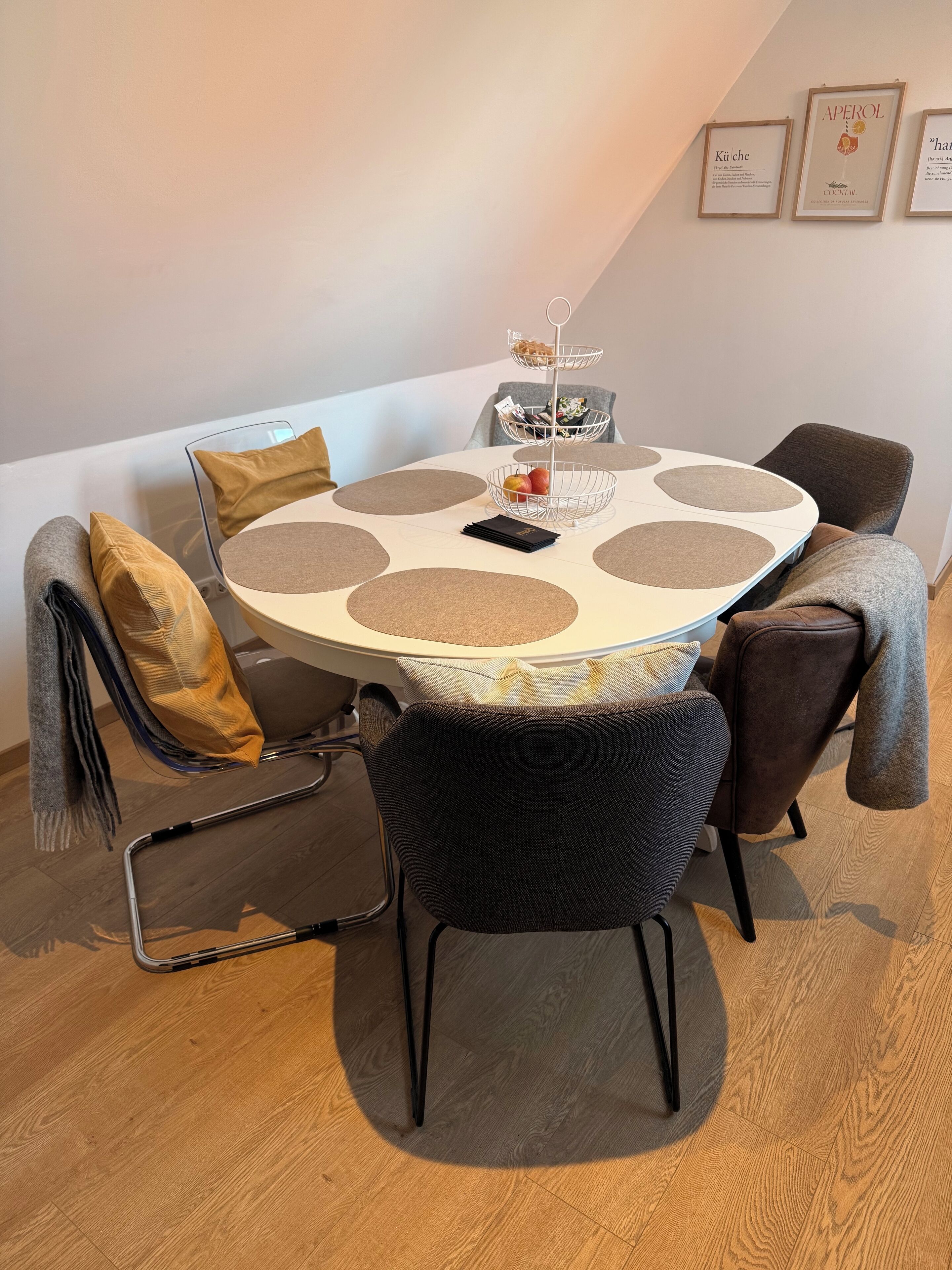 A round table with a white top and brown circular accents, surrounded by four chairs with dark upholstery. A small white stand with a glass top and a few items on top is placed in the center.