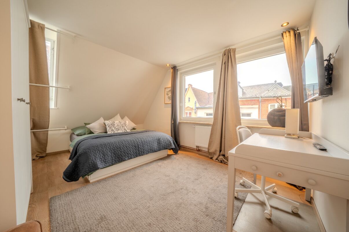 A bedroom with a bed, a desk, a chair, a window with curtains, and a TV on the wall.