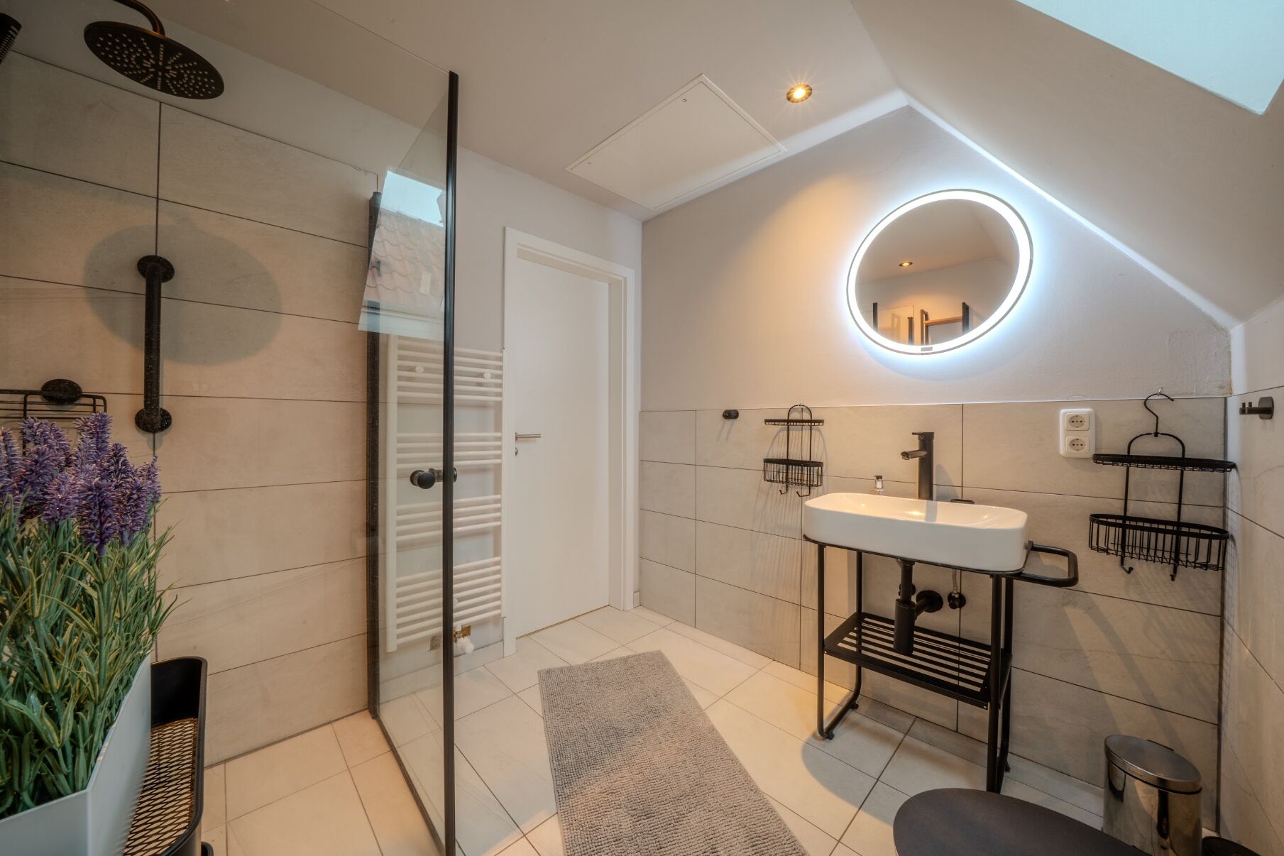 A modern bathroom with a glass shower enclosure, a white sink with a black frame, and a round mirror with a light.