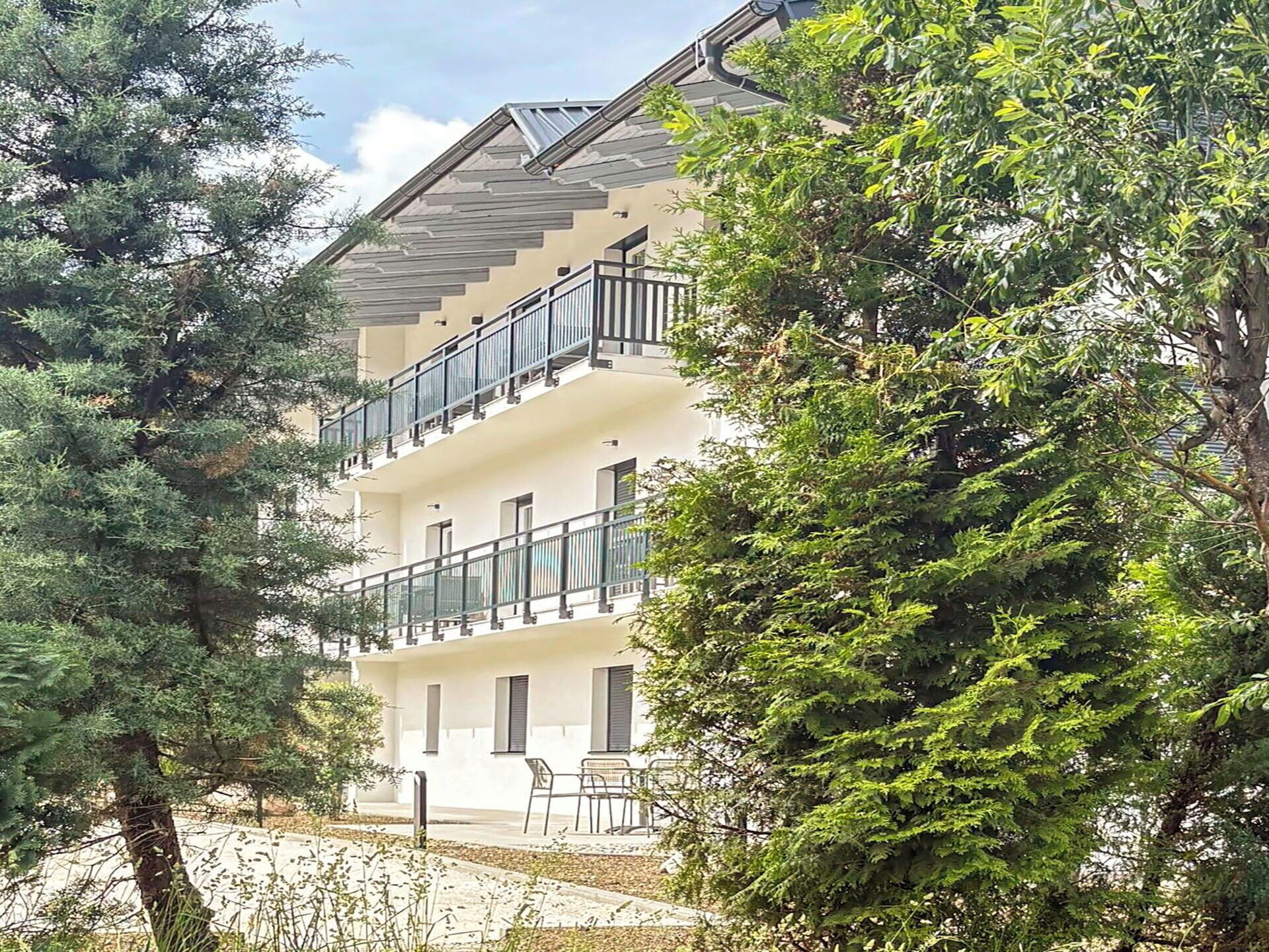 Un immeuble à plusieurs étages doté de balcons, une zone pavée avec des chaises et des arbres tout autour.