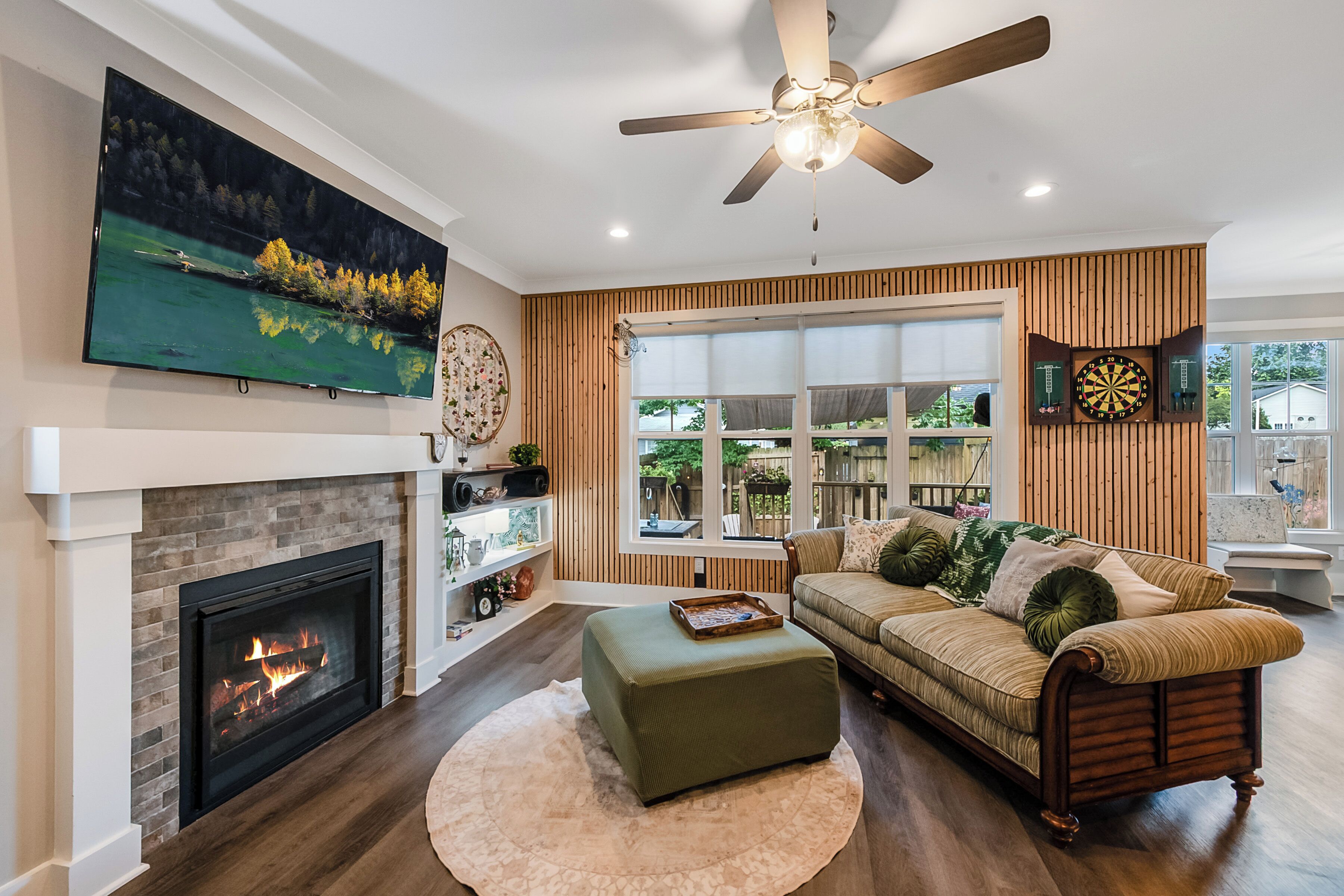 A modern living room with a fireplace, a large flat-screen TV, a ceiling fan, and a large window with a view of the outdoors.