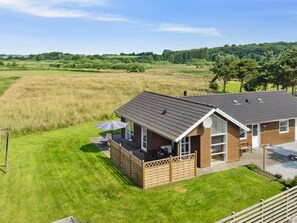 Een houten huis met twee verdiepingen, een balkon, een gazon en uitzicht op een groen veld en bomen.