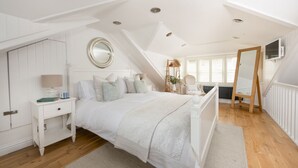 Et soveværelse med skråvæg, seng med hvide sengetøj, et natbord med lampe, et rundt spejl over sengen, og en træstige med en plante.