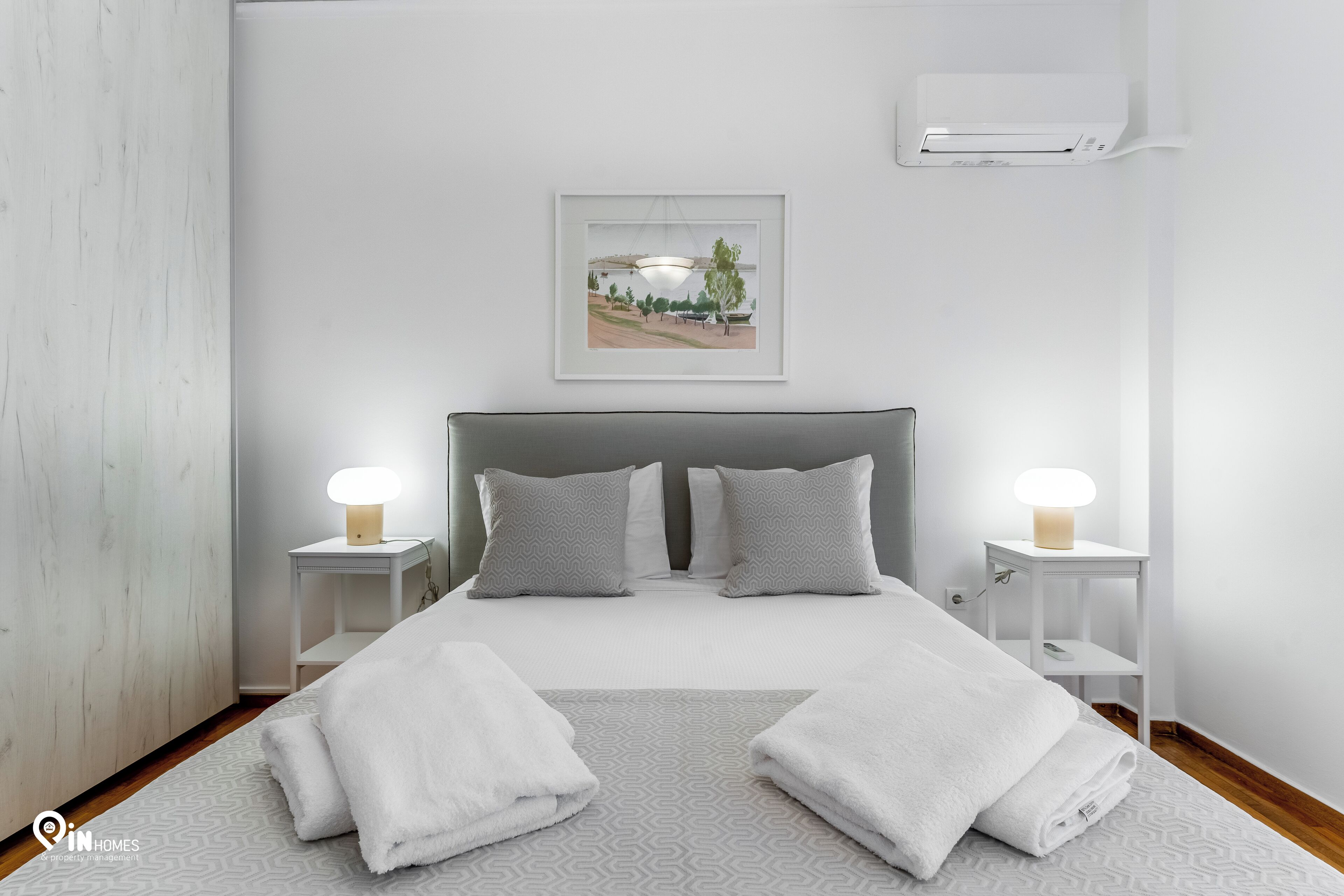 A neatly arranged bedroom with a bed, two bedside tables, a wall-mounted air conditioner, and a framed picture above the bed.