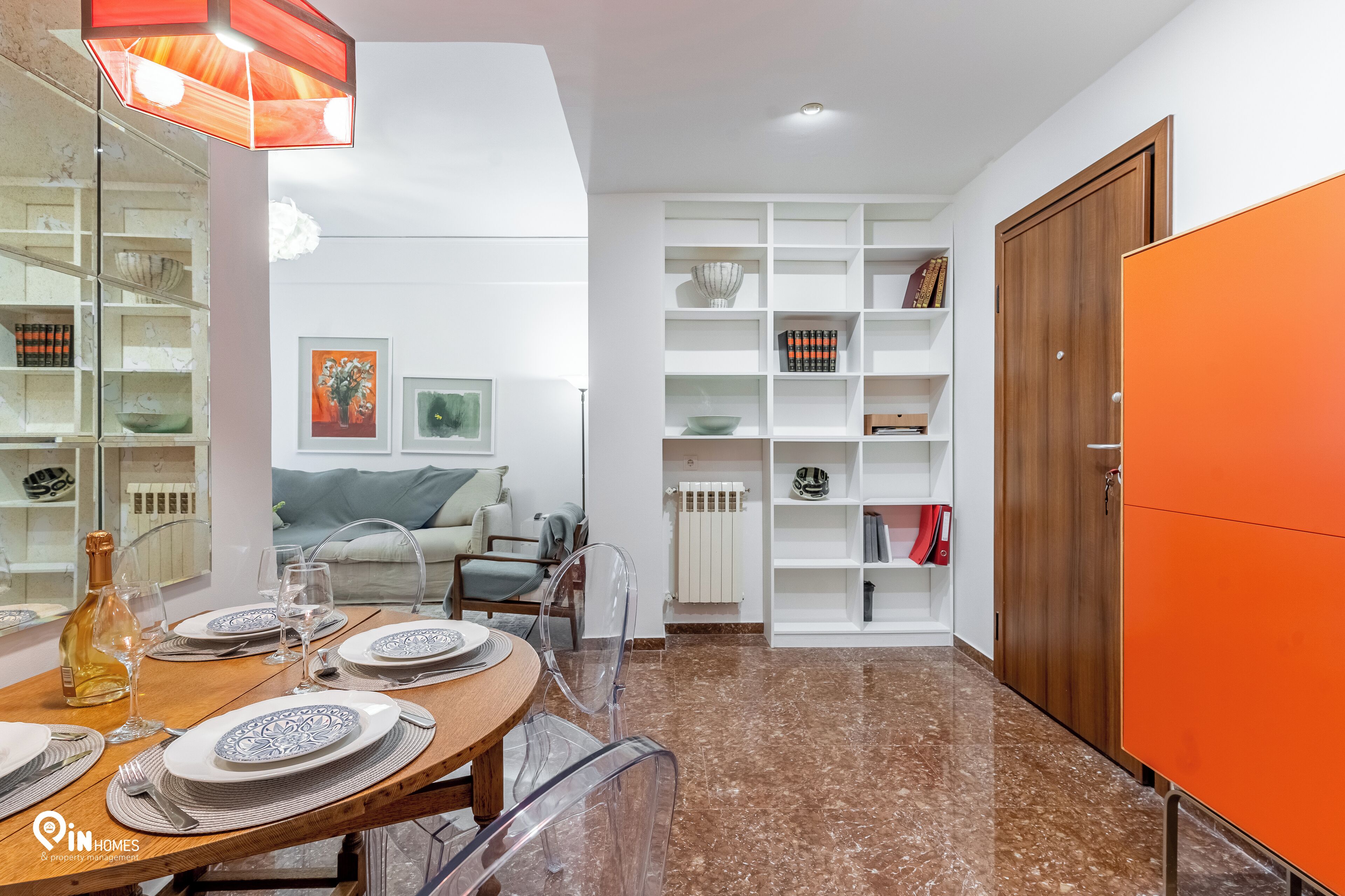 A dining area with a wooden table set for four, clear chairs, and a bottle of wine. There is a white shelving unit with decorative items and books. A wooden wardrobe and a bright orange cabinet are visible.
