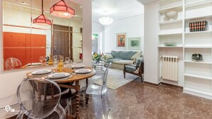 On aperçoit un coin repas avec une table en bois dressée pour quatre personnes, des chaises transparentes et des lampes suspendues. Au fond, on distingue un salon avec un canapé et un radiateur.