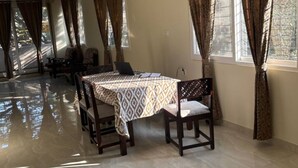 A spacious room with a dining table set for four, large windows with curtains, a ceiling fan, and a tiled floor. - The Yellow Gate - 2BHK - New Apartment - Near Banashankari (Bengaluru)