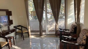A living room with a flat-screen TV, wooden furniture, and a large window with curtains. - The Yellow Gate - 2BHK - New Apartment - Near Banashankari (Bengaluru)