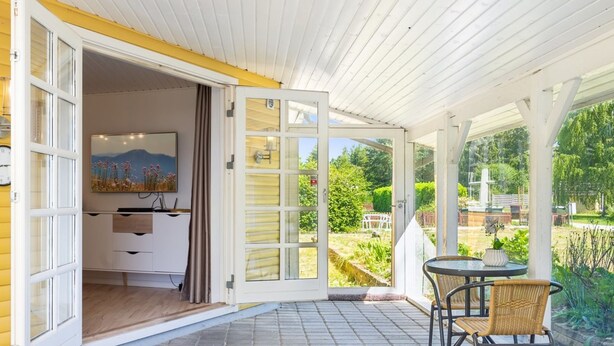 Une terrasse ouverte avec une table et des chaises, un coin cuisine visible à travers une porte ouverte et une vue sur le jardin.