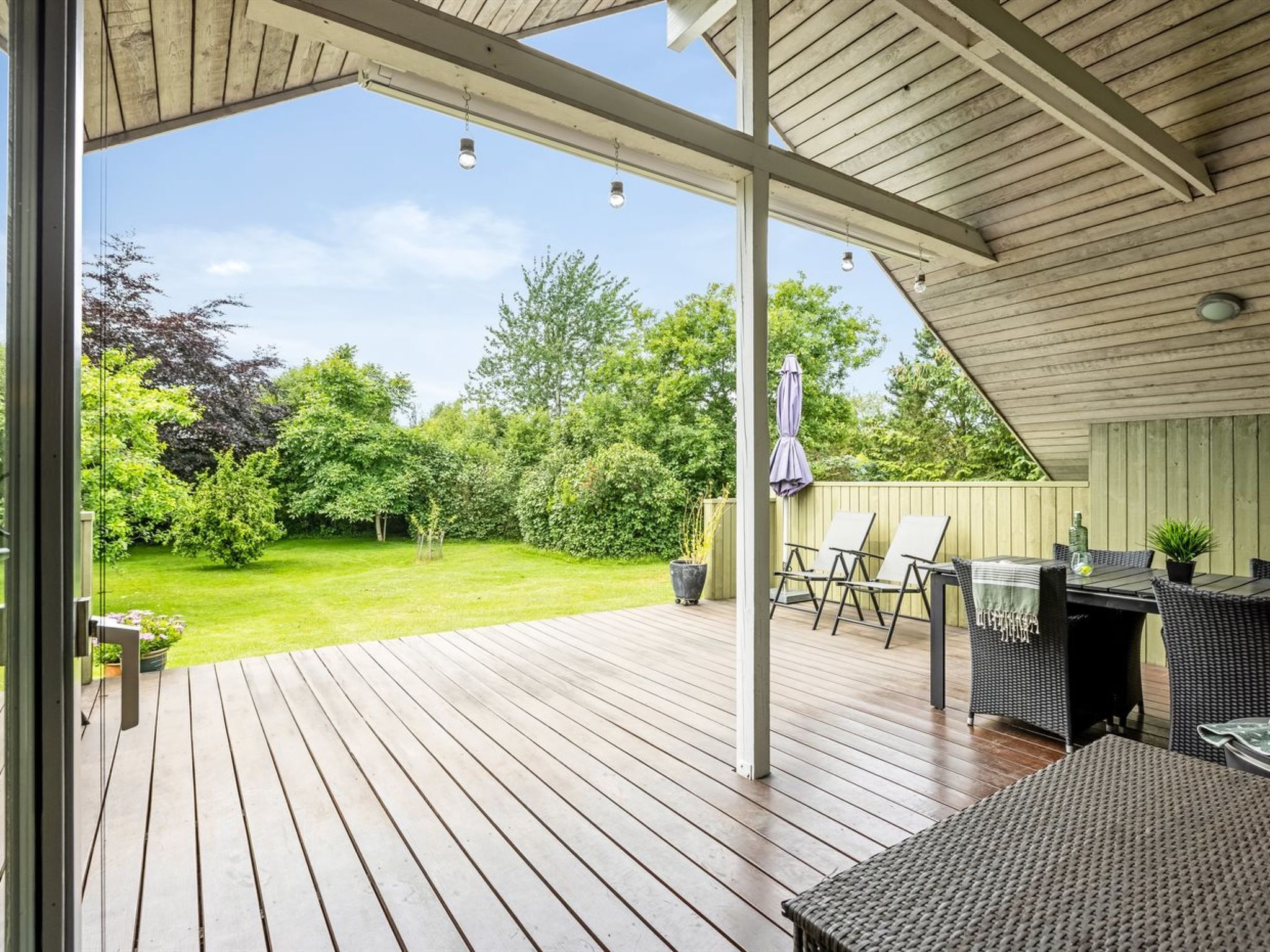 A covered patio with a wooden deck, outdoor furniture, and a view of a lush garden.