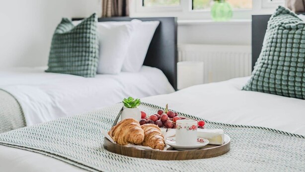 Un lit bien fait, avec un plateau contenant des croissants, des raisins et une tasse de thé, placé devant une fenêtre donnant sur une maison.