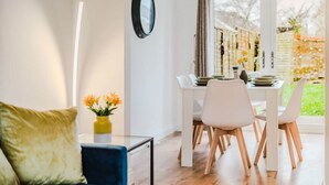 Un coin repas avec une table et des chaises blanches, un vase rempli de fleurs et une vue sur un jardin, visible à travers de grandes fenêtres.