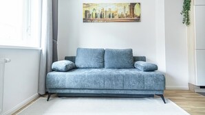Uma sala de estar moderna com um sofá cinzento, uma pintura de paisagem urbana na parede e um armário branco com uma planta.