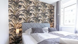 Quarto de hotel com uma janela grande, cama com roupa de cama branca, cabeceira cinza e parede com padrão de folhagem.