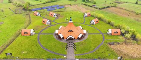 An aerial view of a campsite with multiple dome-shaped tents arranged in a circular pattern around a central building with a conical roof. There are solar panels visible in the background, and a parking area with several cars.