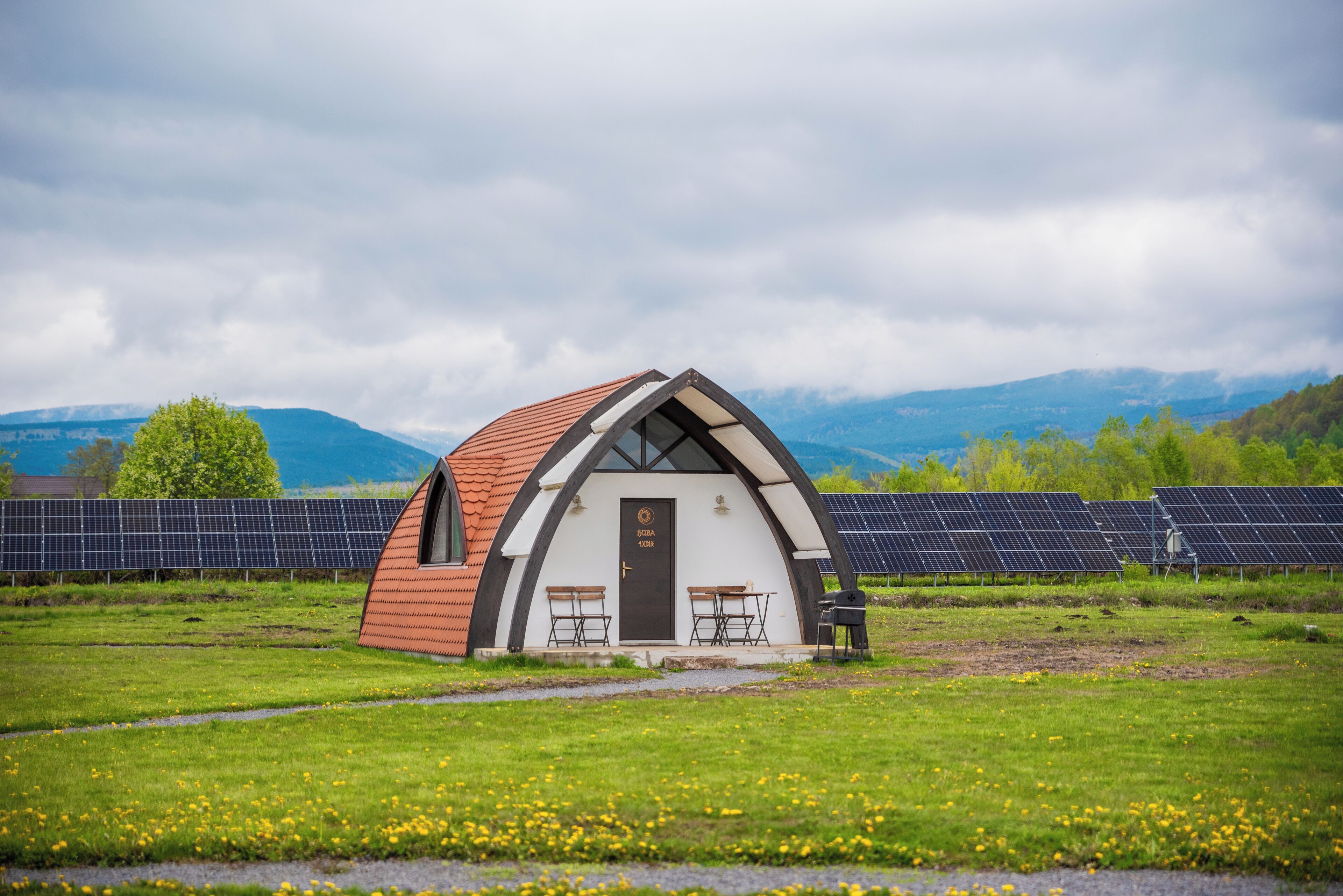 A small, white, arched building with a red roof, two tables, and a door labeled "SOBRA". It is surrounded by a grassy field and solar panels in the background.
