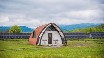 A small, white, arched building with a red roof, two tables, and a door labeled "SOBRA". It is surrounded by a grassy field and solar panels in the background.
