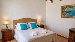 A neatly made bed with white and blue pillows, two bedside lamps, a wooden nightstand, and a painting of a coastal scene on the wall. - Authentic Menorcan country house, close to the capital and beaches, in total calm. (Llucmaçanes)