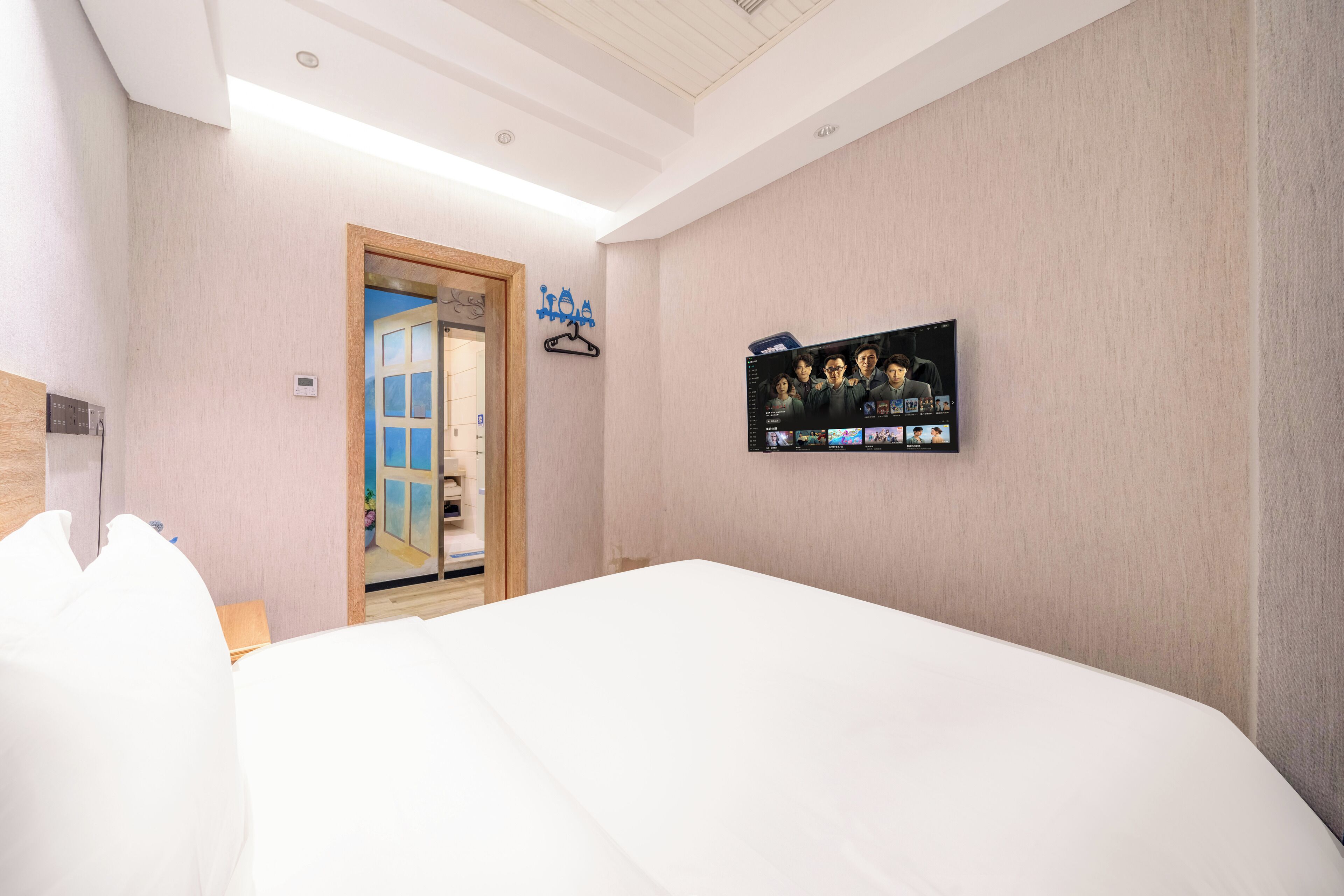 Een hotelkamer met een groot bed, een aan de muur bevestigde televisie waarop een videogesprek met een groep mensen te zien is, een houten deur en een klein plankje met een blauwe kleerhanger.