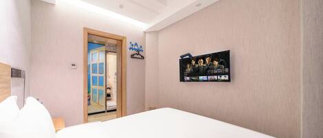 Een hotelkamer met een groot bed, een aan de muur bevestigde televisie waarop een videogesprek met een groep mensen te zien is, een houten deur en een klein plankje met een blauwe kleerhanger.
